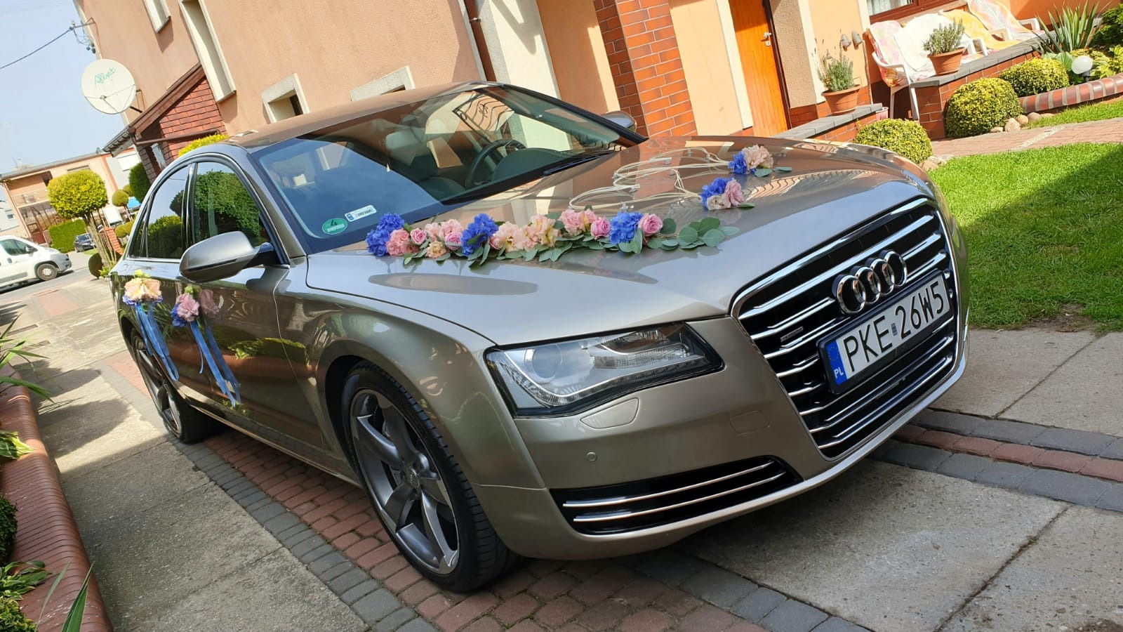 Złoty samochód Audi A8 udekorowany kwiatami i wstążkami, zaparkowany na podjeździe przed domem.