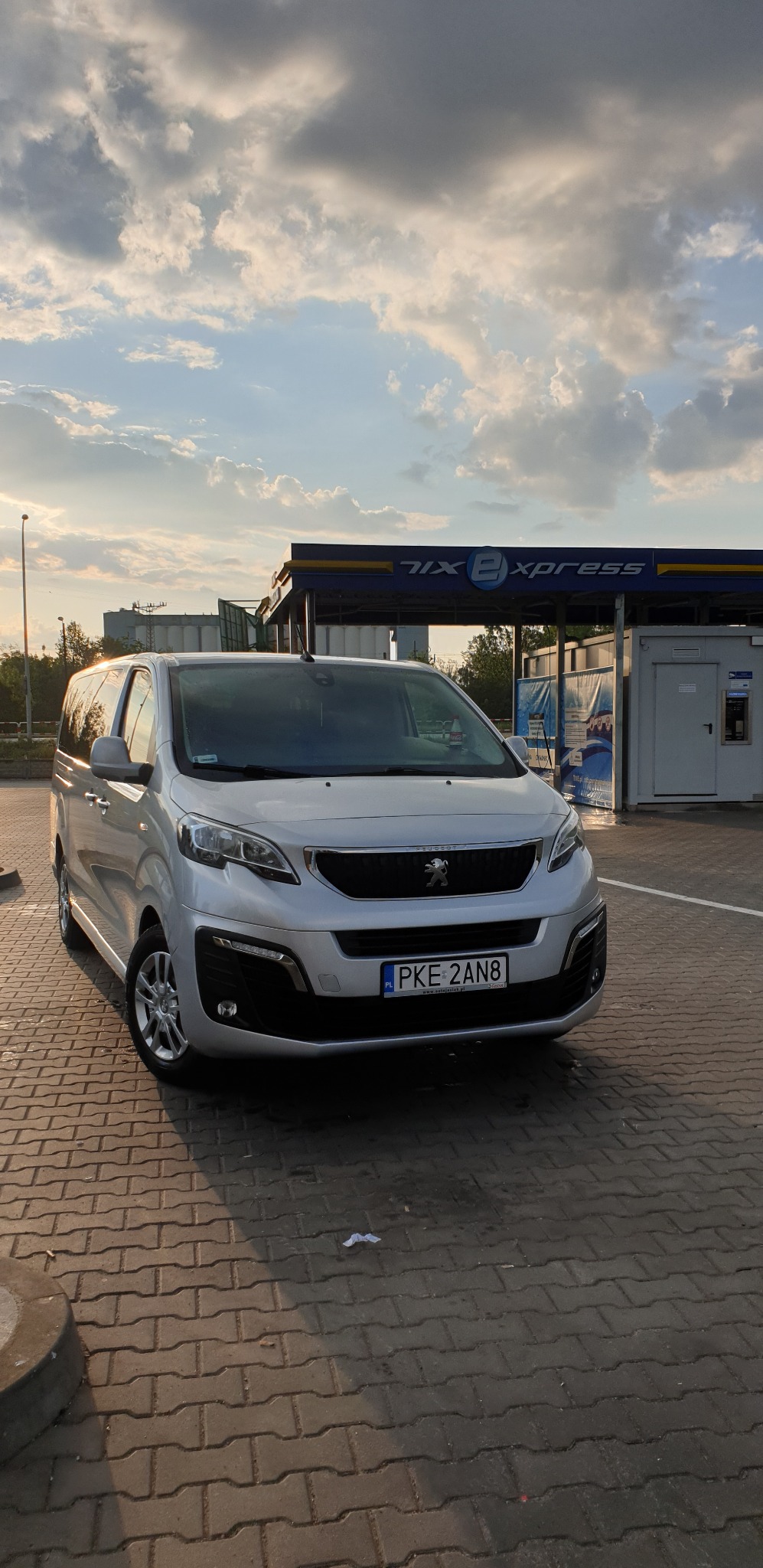 Srebrny Peugeot Expert Tepee zaparkowany przed myjnią samochodową z błękitnym niebem w tle, widoczna tablica rejestracyjna PKE 2AN8.