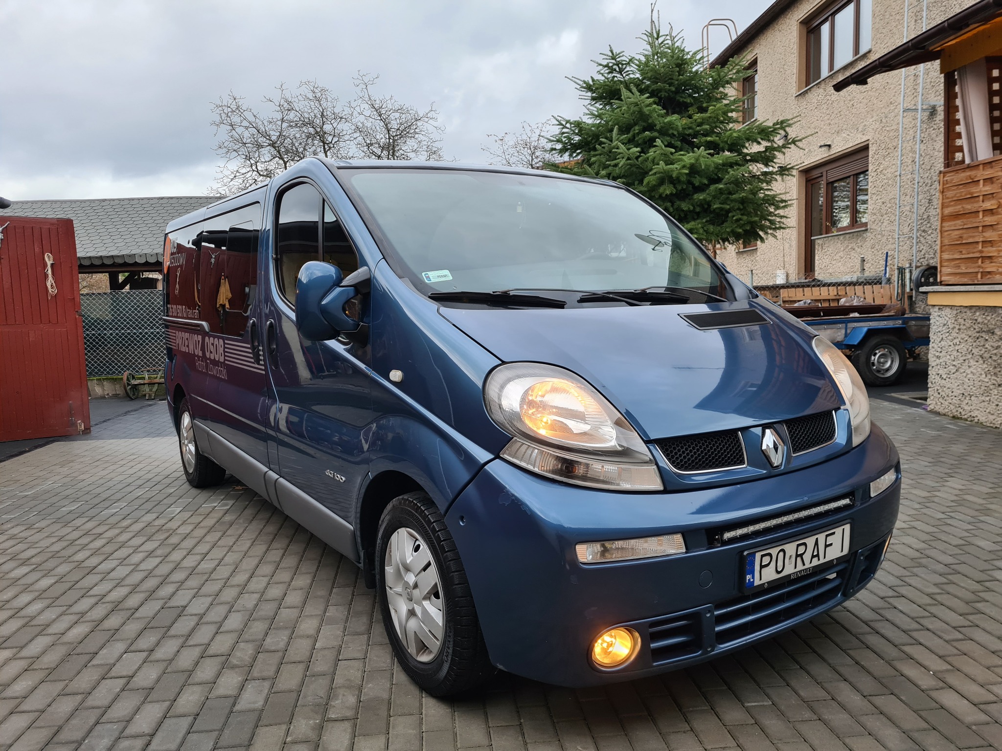 Niebieski bus Renault z napisem 'Przewóz osób' zaparkowany na brukowanej nawierzchni w Kępnie, z włączonymi światłami mijania i przeciwmgielnymi.