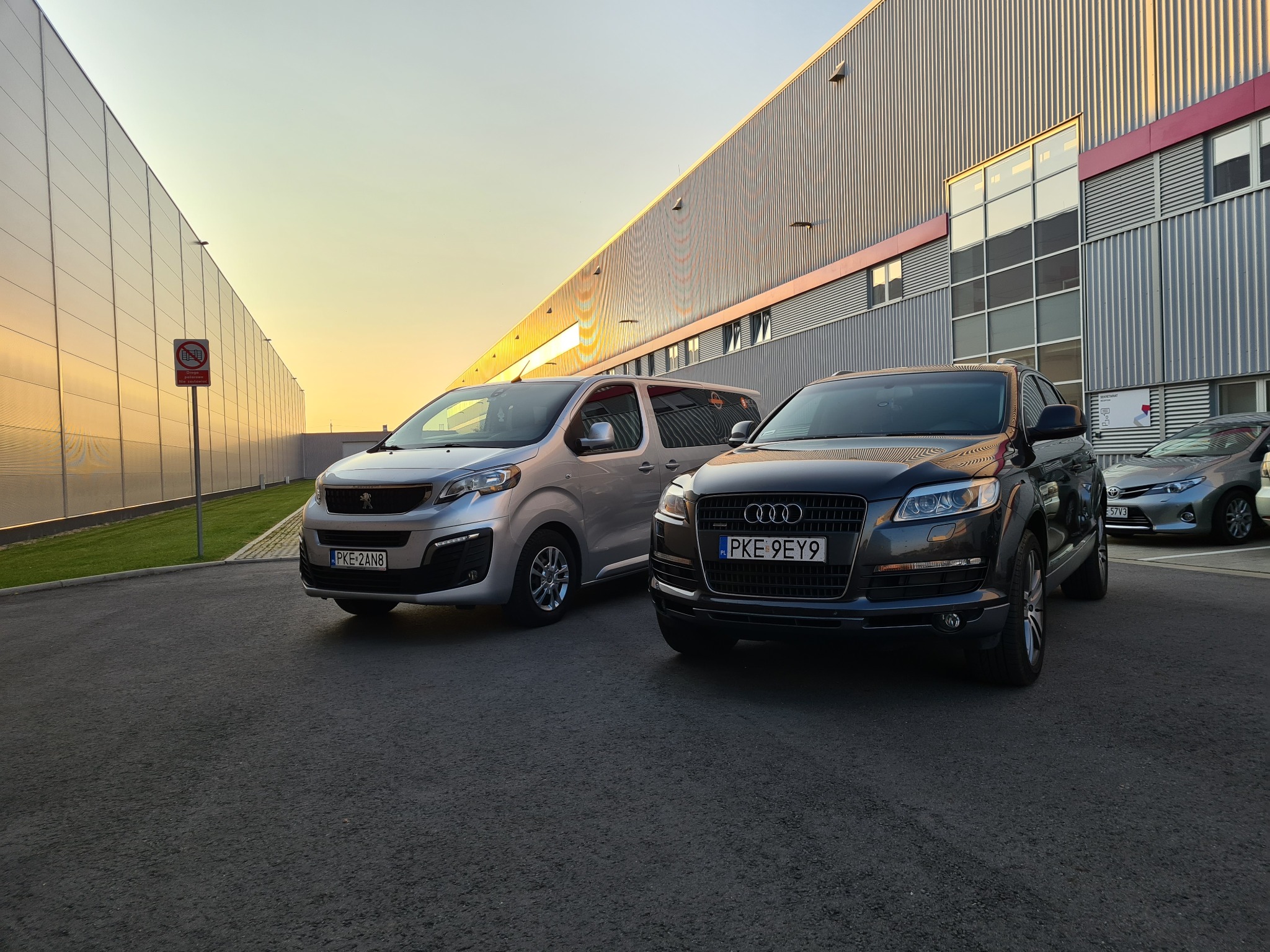 Srebrny Peugeot Expert i ciemny SUV Audi Q7 zaparkowane przed nowoczesnym budynkiem przemysłowym w promieniach zachodzącego słońca, widoczny znak zakazu w tle.