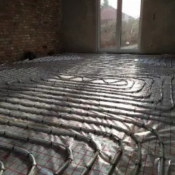 Rozłożona instalacja ogrzewania podłogowego z rur w szarym kolorze, zamocowanych na folii z czerwoną kratką, w surowym wnętrzu z oknem i fragmentem ściany z cegły.