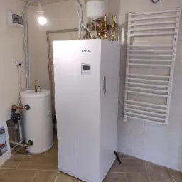 Biała pompa ciepła Kaisai z podłączonym zbiornikiem akumulacyjnym i grzejnikiem drabinkowym w łazience wyłożonej kafelkami.