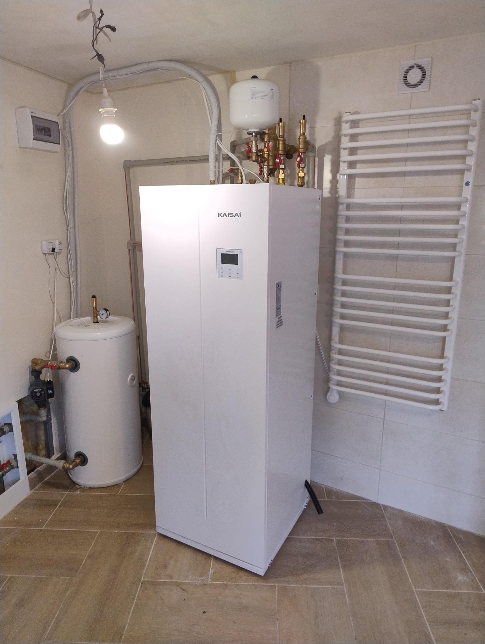 Biała pompa ciepła Kaisai z podłączonym zbiornikiem akumulacyjnym i grzejnikiem drabinkowym w łazience wyłożonej kafelkami.
