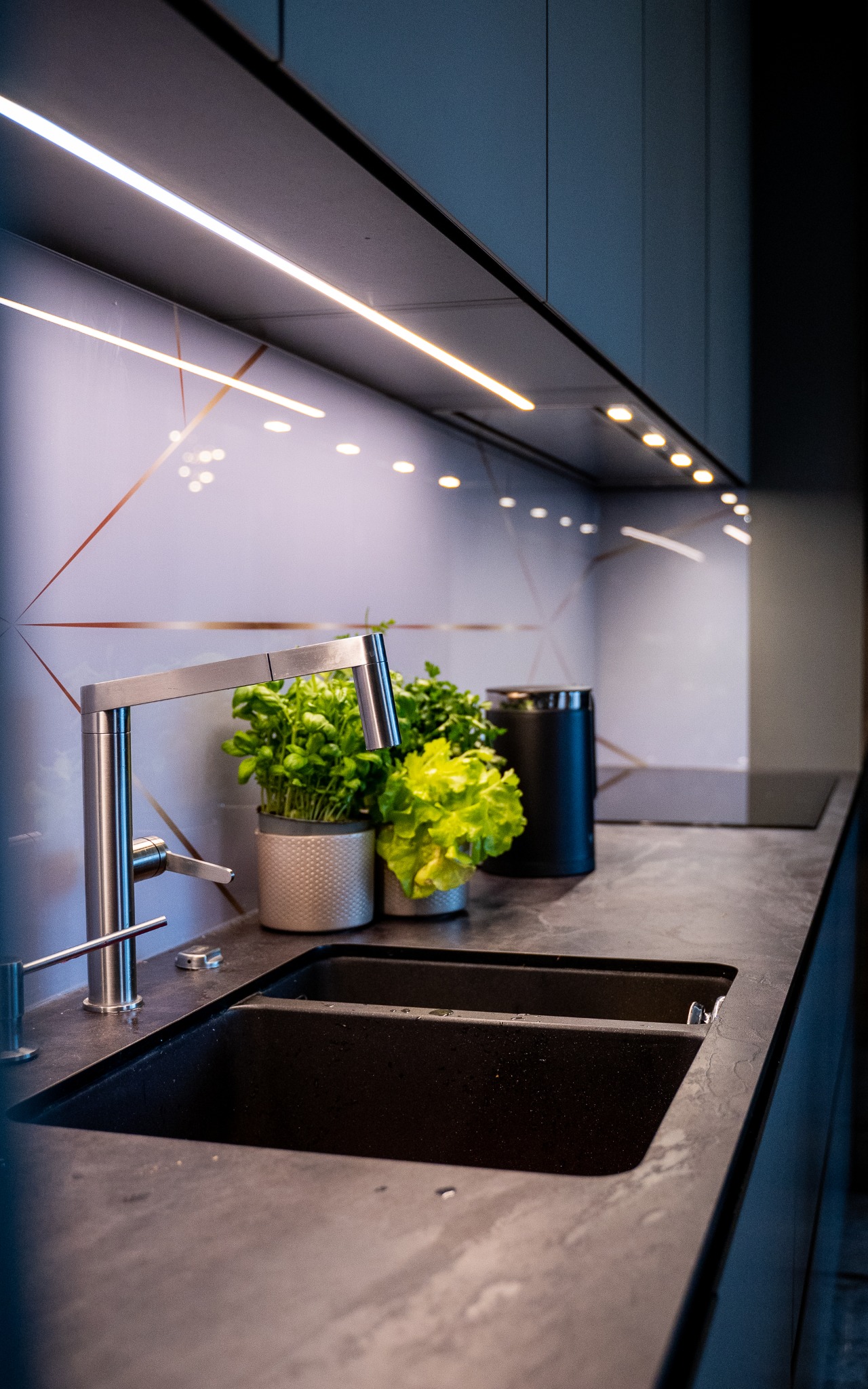 Nowoczesna, ciemna kuchnia z podświetlanym blatem, dwukomorowym zlewozmywakiem, minimalistyczną baterią i roślinami w doniczkach na tle ściany z geometrycznym wzorem.
