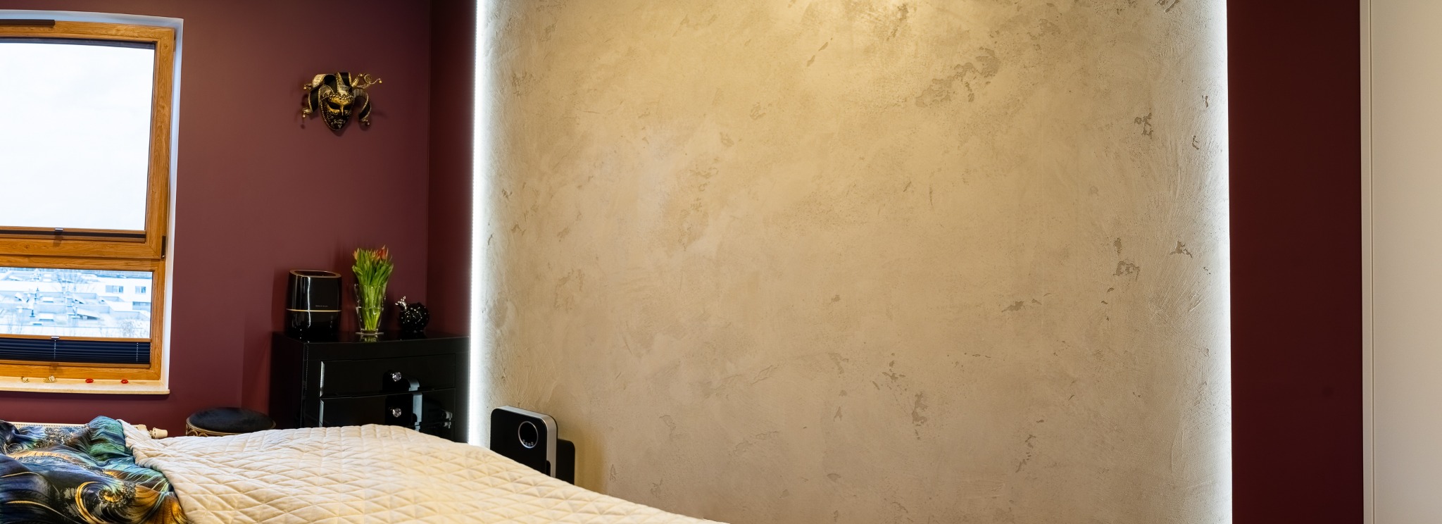 Sypialnia z beżową ścianą o fakturze betonu podświetloną pionowym paskiem LED, burgundowa ściana z dekoracyjną maską wenecką, czarna komoda z kwiatami w wazonie i widokiem na miasto za oknem.