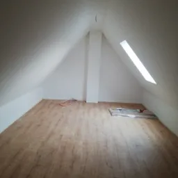 Poddasze z ułożonymi panelami podłogowymi, widoczne kable elektryczne przy ścianie i zapakowane panele w folii. Ściany pomalowane na biało, okno dachowe wpuszczające jasne światło.