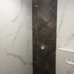 Ściana łazienki wyłożona białymi i ciemnobrązowymi płytkami imitującymi marmur, widoczny otwór na baterię w ciemnej płytce oraz zabudowa stelaża podtynkowego WC w białej płytce.