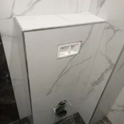 Stelaż podtynkowy WC obudowany płytkami imitującymi marmur, widoczne przyłącze kanalizacyjne i otwór na przyciski spłukiwania.