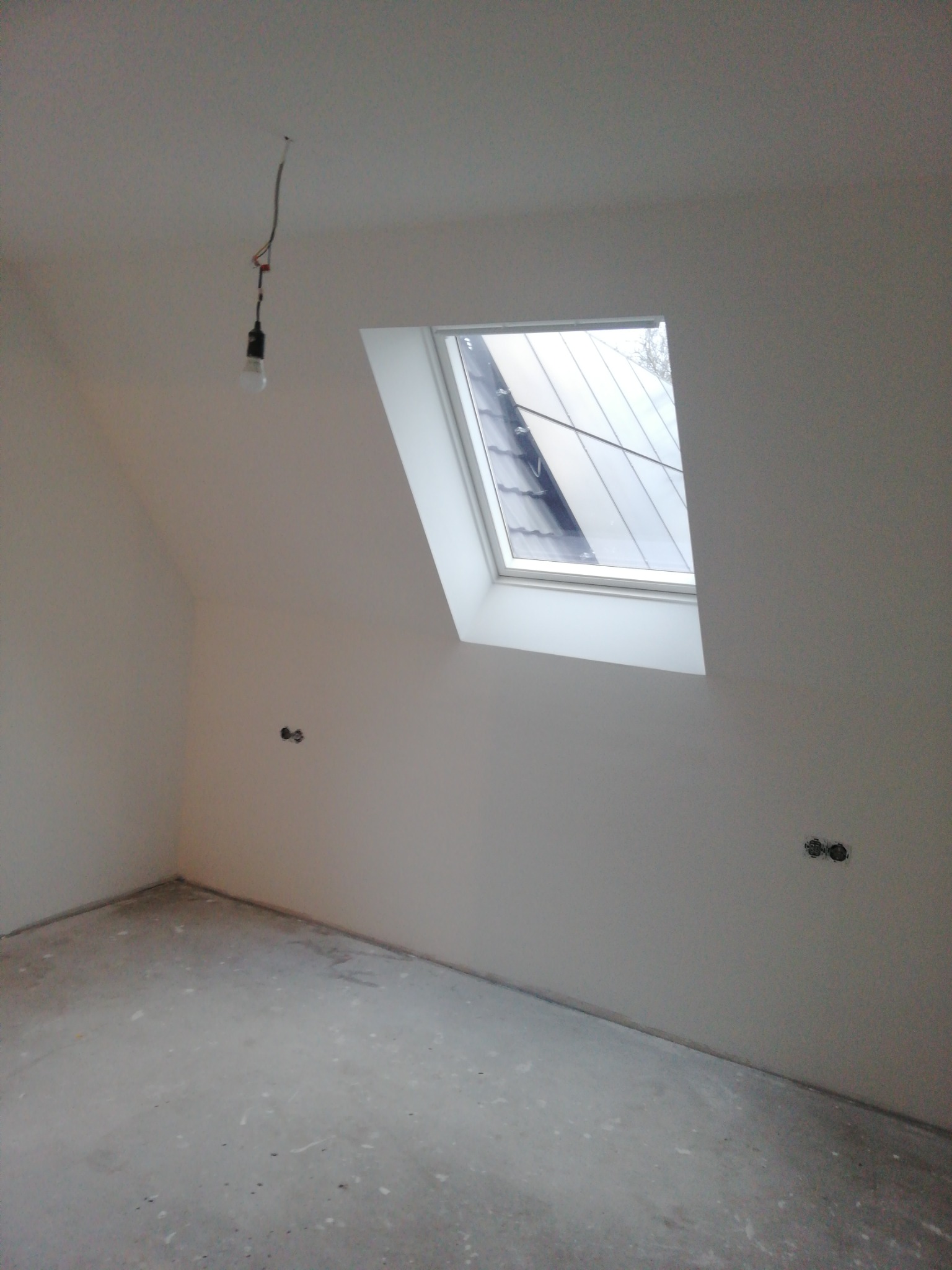 Poddasze w trakcie prac przygotowawczych: surowe ściany po szpachlowaniu, betonowa podłoga, okno dachowe z widokiem na dach, wisząca żarówka na kablu, gniazdka elektryczne.