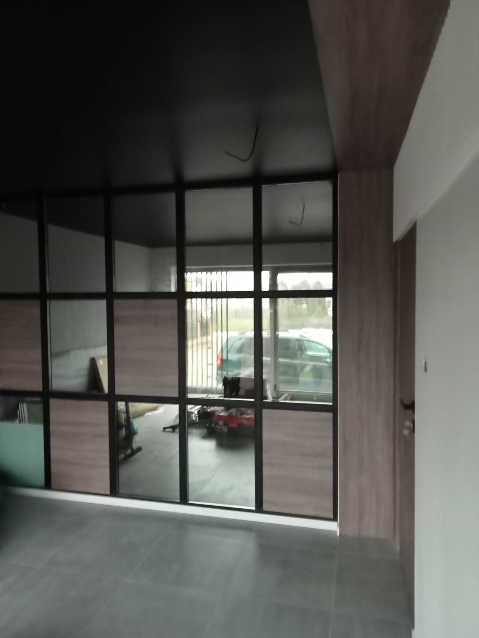 Nowoczesna szklana ścianka działowa z drewnianymi panelami w ramie z czarnego metalu oddzielająca pomieszczenie od garażu, szara podłoga z płytek.