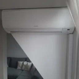 Biały klimatyzator marki Innova zamontowany na ścianie w rogu pokoju, widoczny fragment szarej sofy z poduszkami w tle.