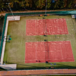 Widok z góry na dwa korty tenisowe z czerwoną nawierzchnią i zielonym otoczeniem, obok plac zabaw dla dzieci z zielonymi elementami, całość otoczona zieloną siatką i drzewami.