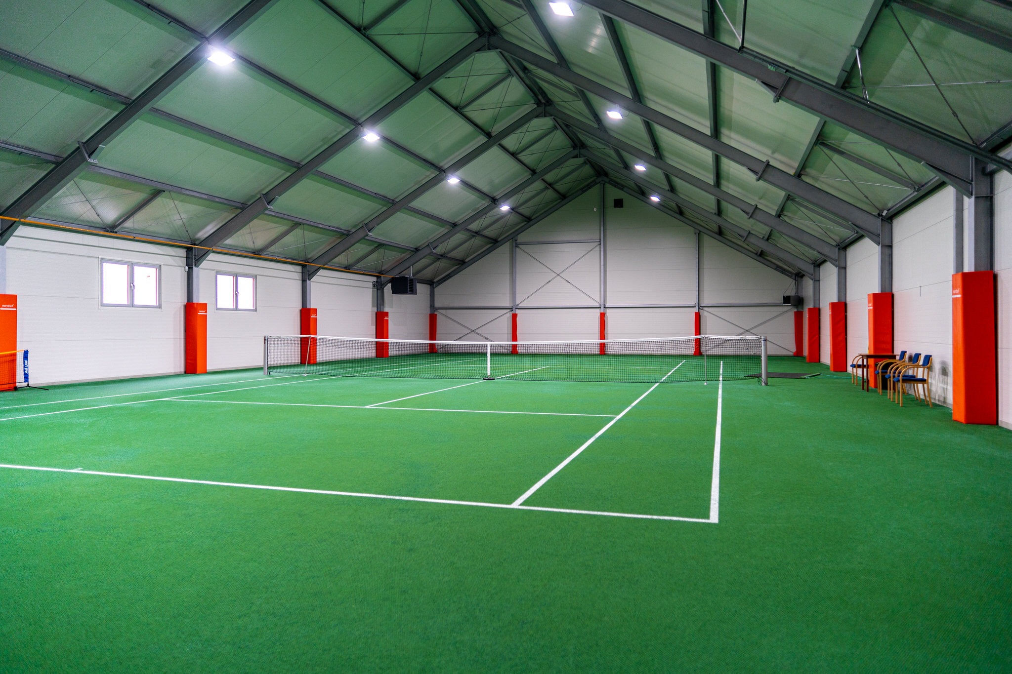 Wnętrze hali sportowej z zieloną nawierzchnią kortu tenisowego, białe linie wyznaczające pole gry, siatka, metalowa konstrukcja dachu z oświetleniem, pomarańczowe słupy ochronne przy ścianach, okna...