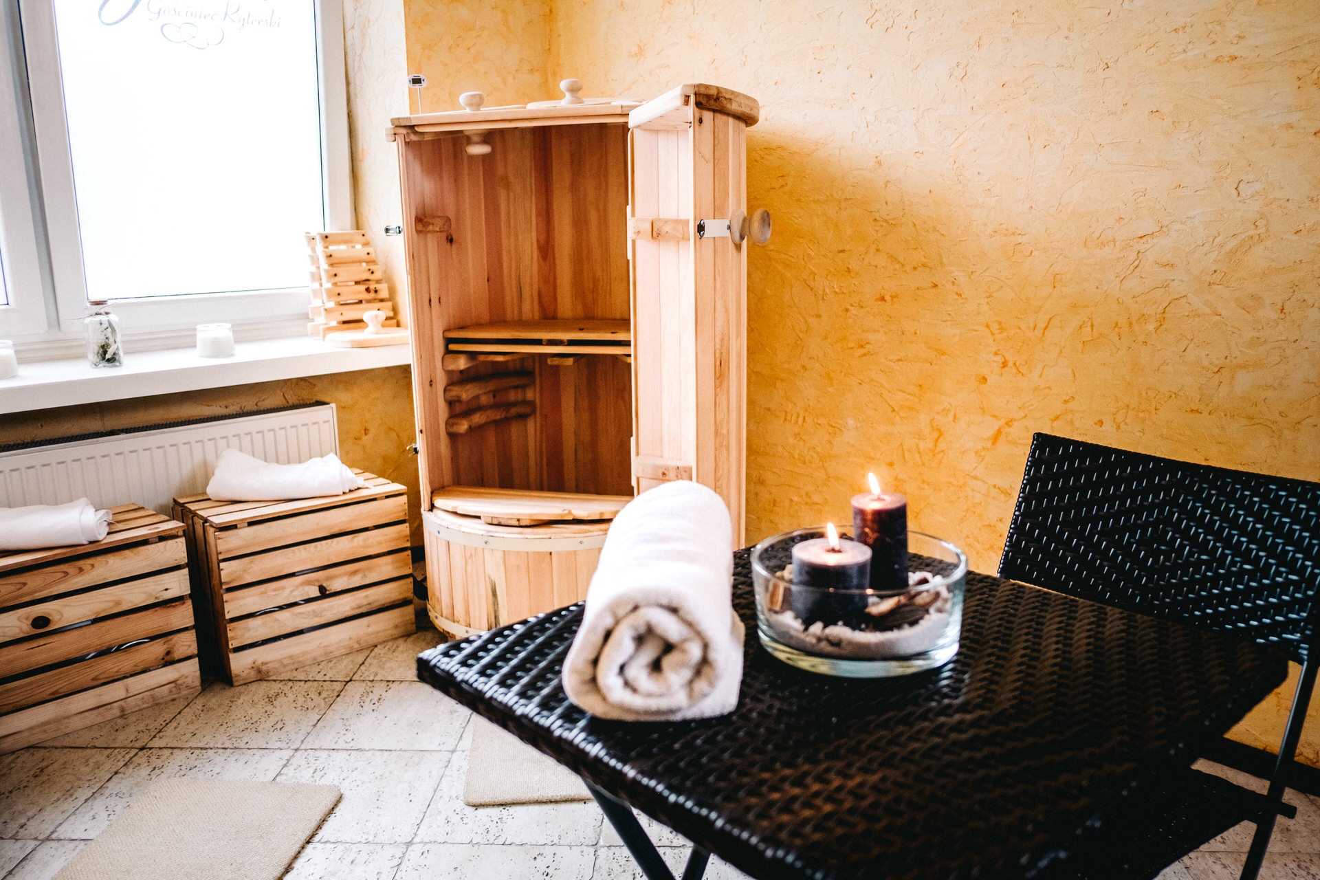 Drewniana sauna parowa z otwartymi drzwiami, zwinięty biały ręcznik i zapalone świece na czarnym stoliku w pomieszczeniu z pomarańczowymi ścianami.