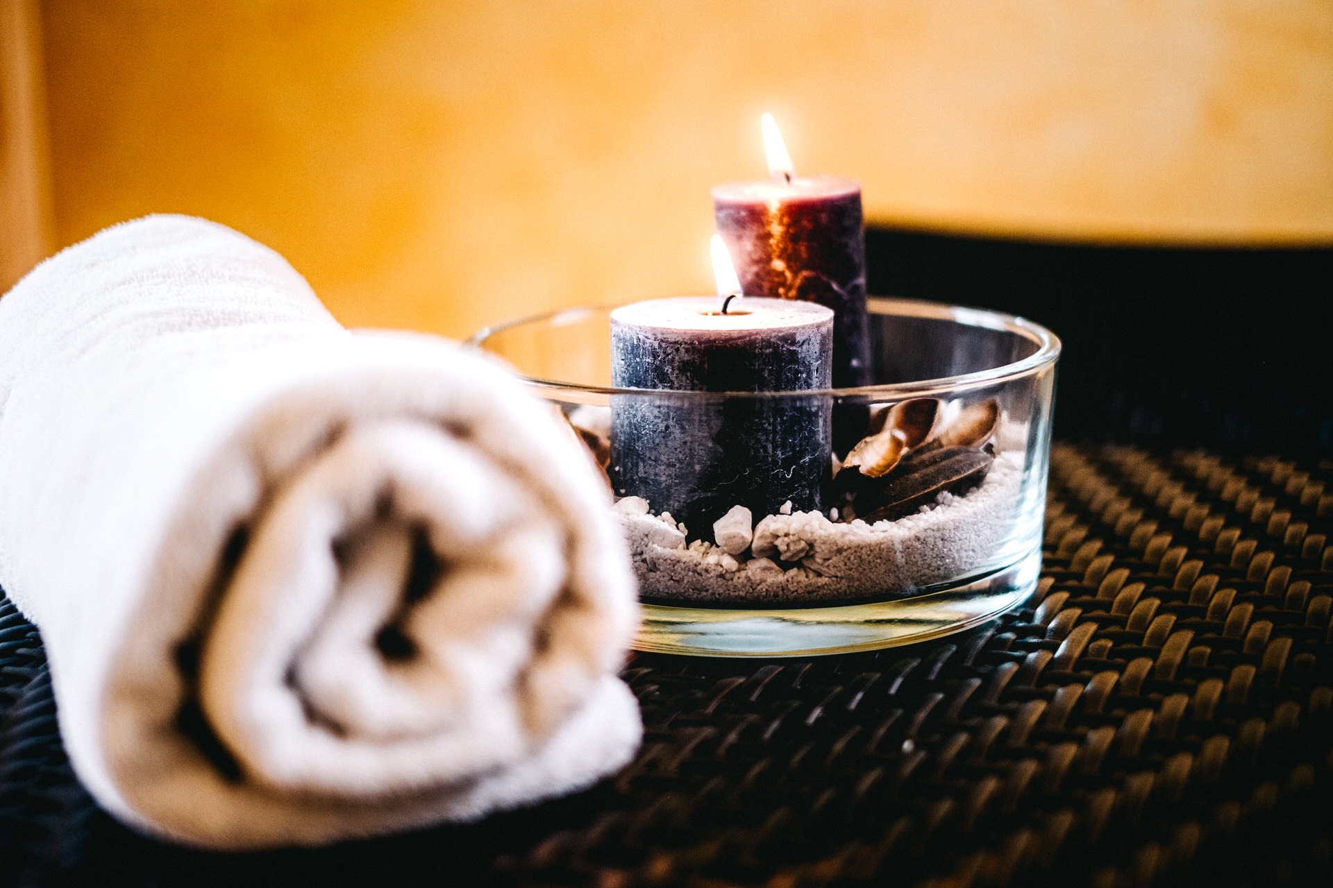 Dwie zapalone świece w szklanym naczyniu z piaskiem i kamieniami, obok zwinięty biały ręcznik na rattanowym stoliku.