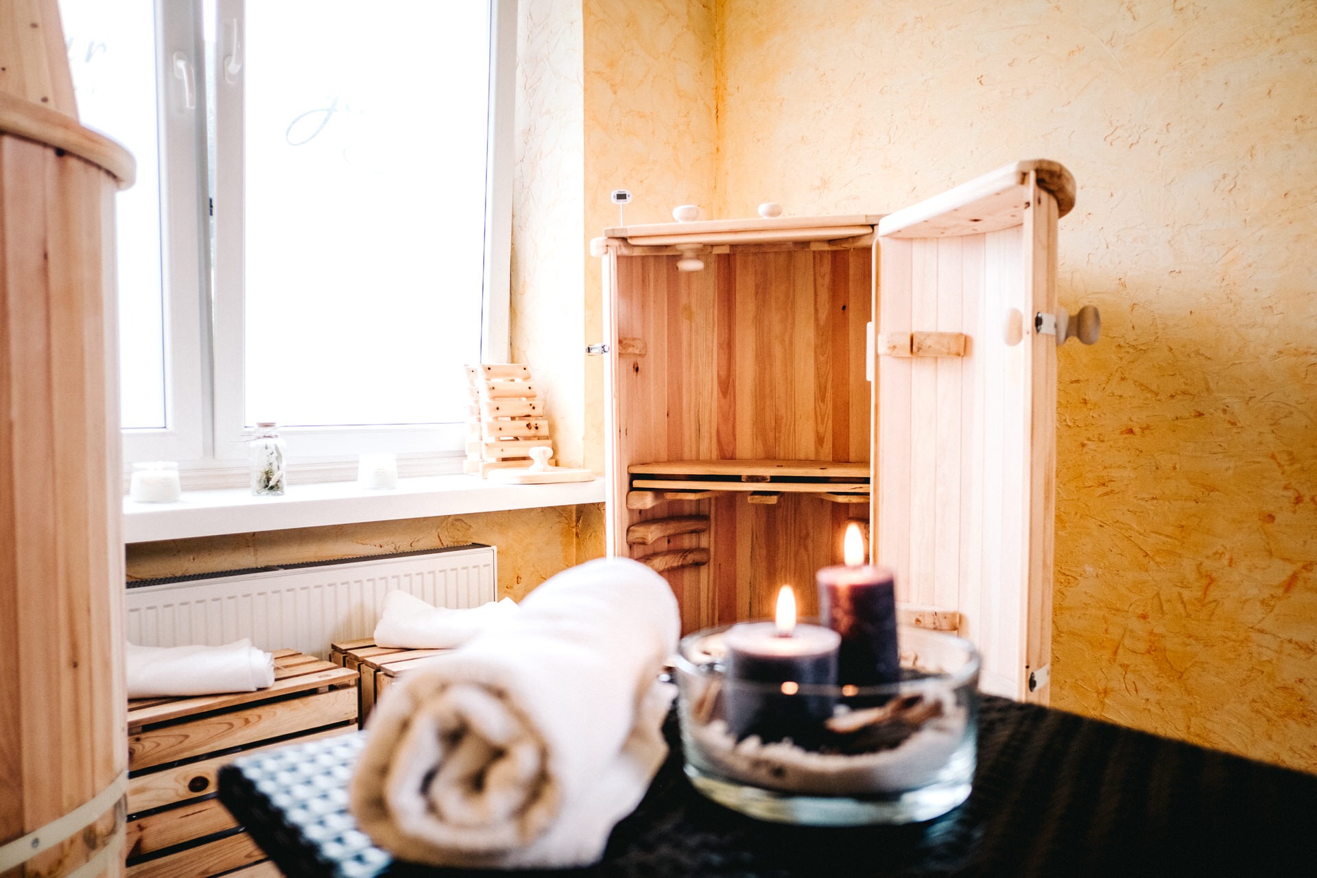 Drewniana sauna z otwartymi drzwiami, obok ręczniki, świece w szklanym naczyniu i okrągła beczka, tworzące relaksującą atmosferę SPA.