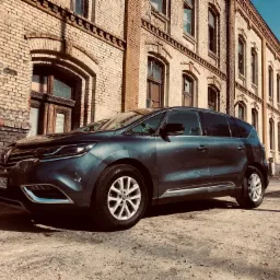 Ciemny minivan zaparkowany przed ceglanym budynkiem z wyraźnymi śladami zużycia, widoczne detale karoserii i felgi.