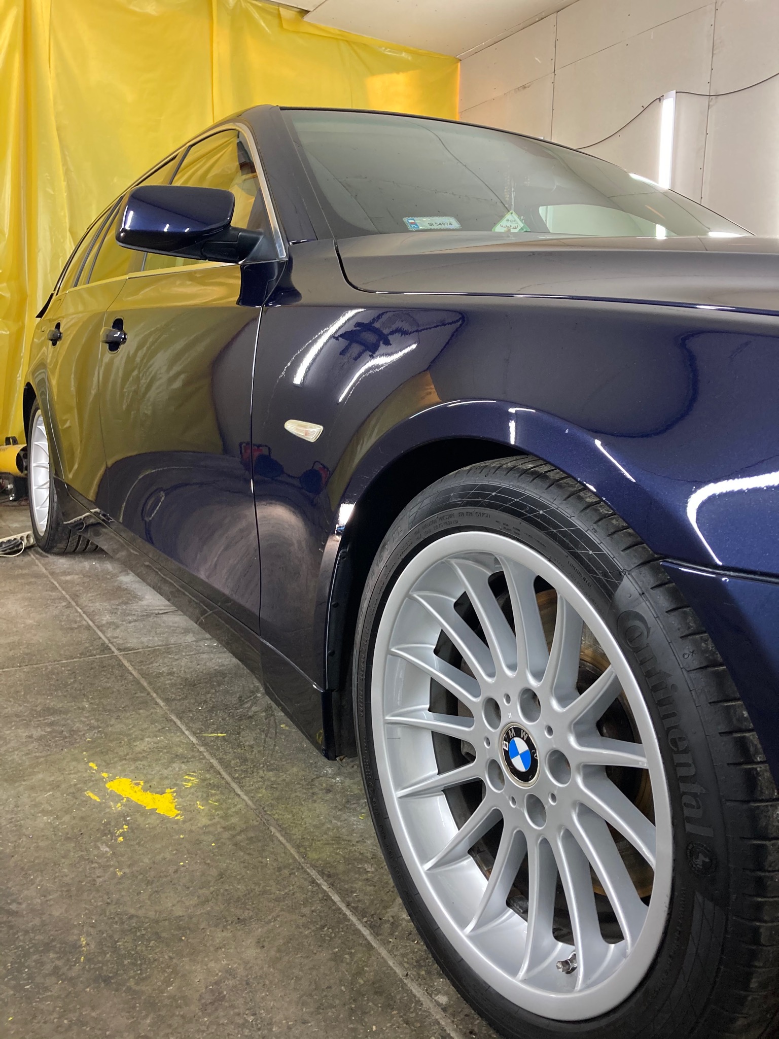 Błyszczący, granatowy samochód kombi marki BMW, sfotografowany pod kątem w warsztacie z żółtymi zasłonami w tle, po umyciu, z widocznymi kroplami wody na lakierze.