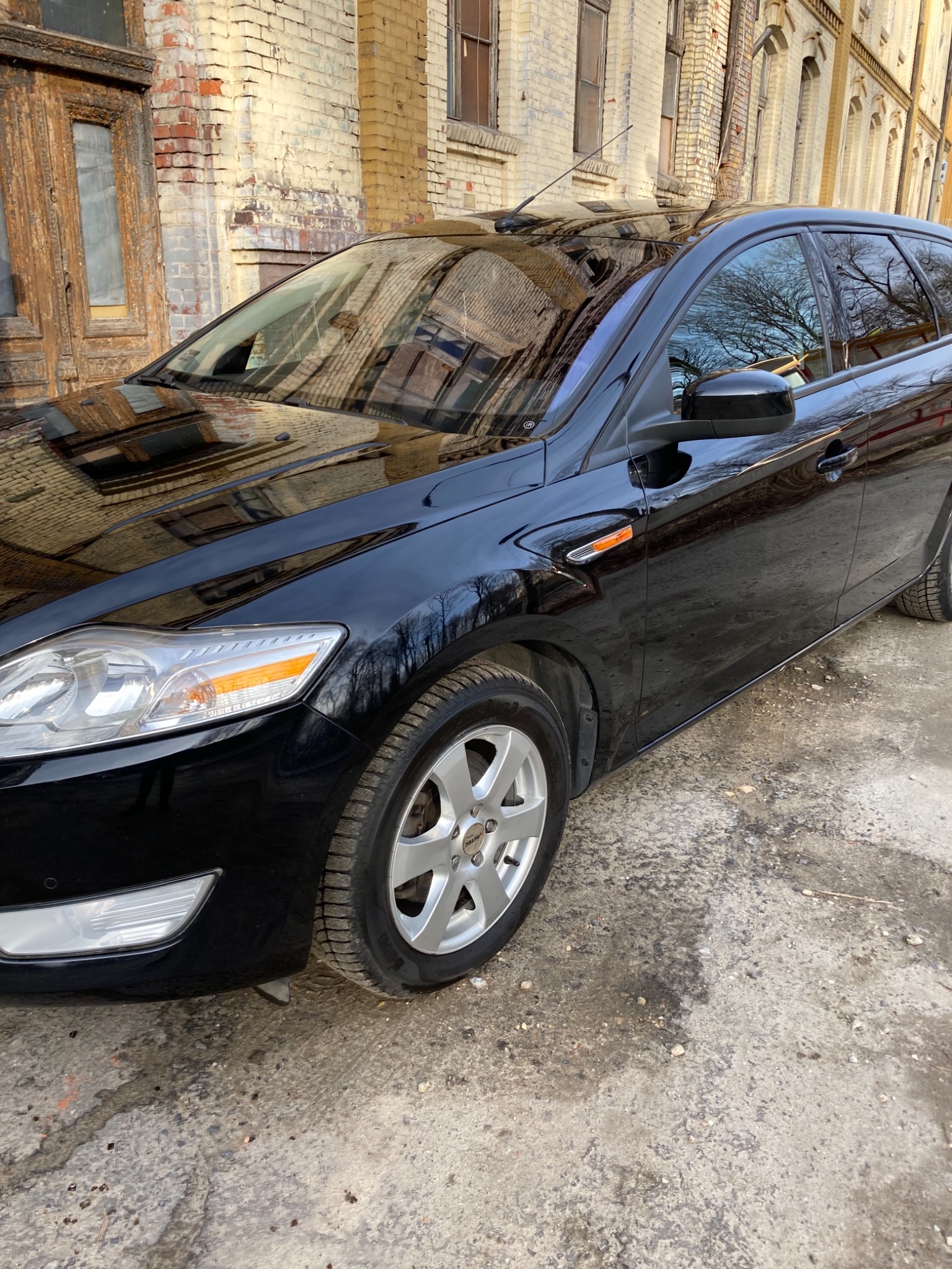 Błyszczący, czarny kombi zaparkowany na zewnątrz budynku z cegły, odbicie drzew w szybach, widoczne aluminiowe felgi i przedni reflektor.