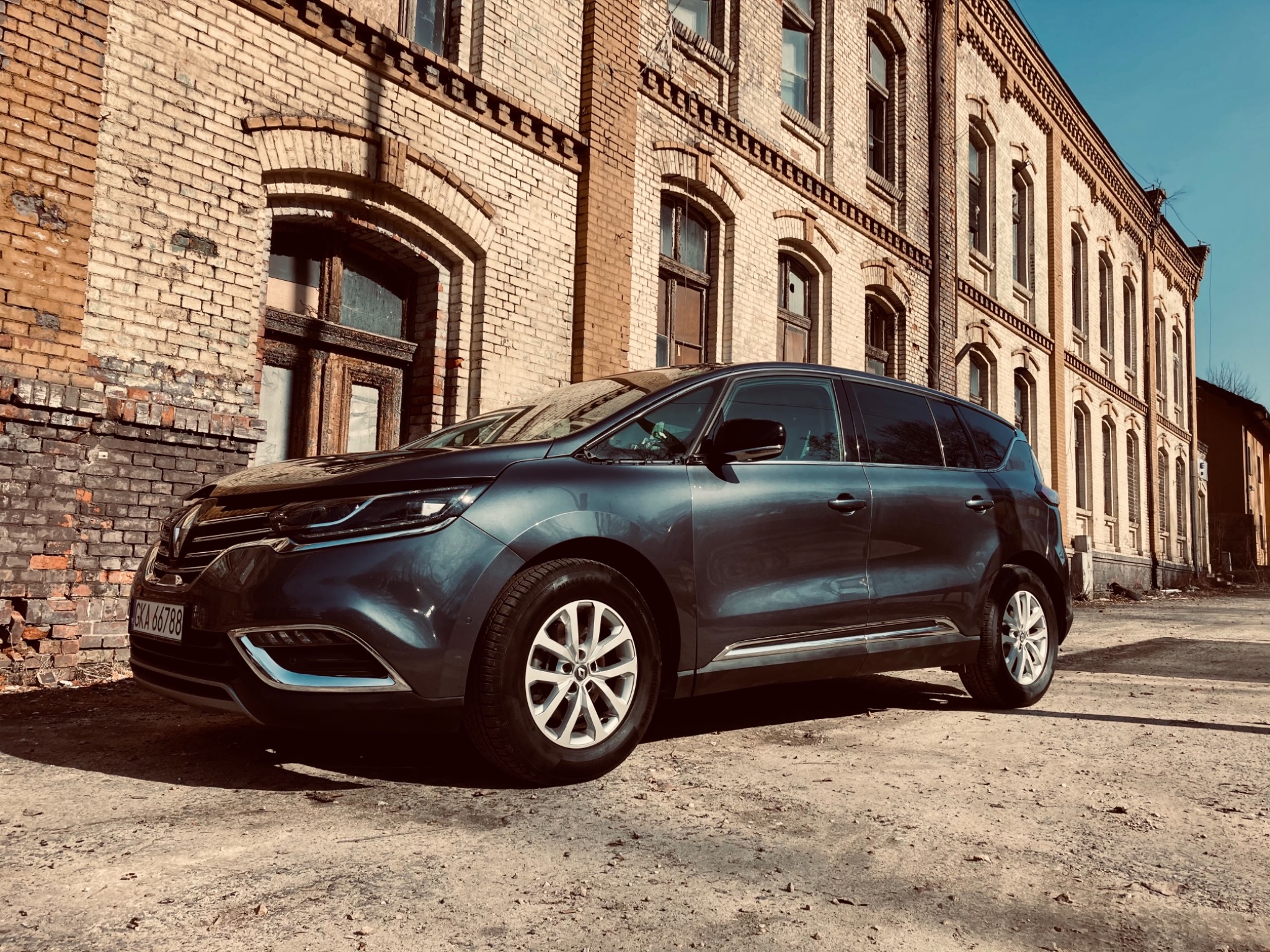 Ciemny minivan zaparkowany przed ceglanym budynkiem z wyraźnymi śladami zużycia, widoczne detale karoserii i felgi.