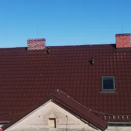 Ciemnobrązowy dach z dachówki trapezowej z dwoma ceglanymi kominami i oknami dachowymi, widok z lekkiego ukosa z góry w słoneczny dzień.