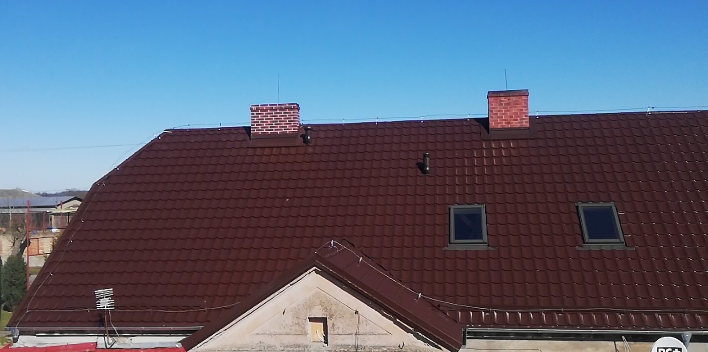 Ciemnobrązowy dach z dachówki trapezowej z dwoma ceglanymi kominami i oknami dachowymi, widok z lekkiego ukosa z góry w słoneczny dzień.