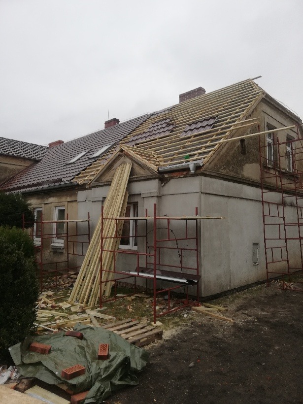 Renowacja dachu na starym domu: częściowo ułożona dachówka, odsłonięta więźba, rusztowania, deski opierające się o ścianę, gruz i cegły na plandece.