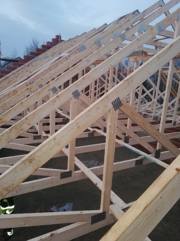 Konstrukcja drewniana dachu w trakcie montażu, widoczne elementy więźby dachowej połączone metalowymi płytkami, w tle mur z czerwonej cegły i pochmurne niebo.