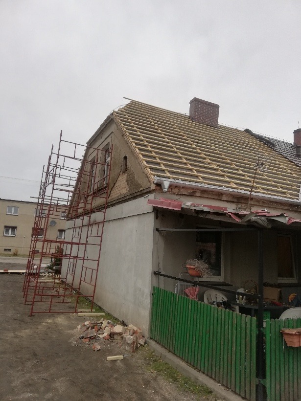 Dom w trakcie remontu dachu, widoczne rusztowanie, stara więźba i komin z cegły, fragment elewacji z ubytkiem tynku.