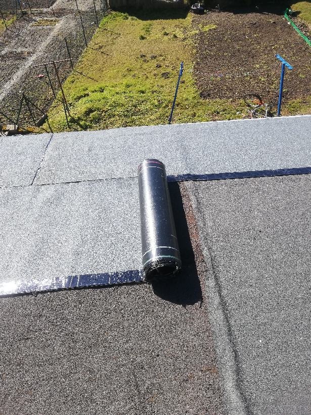 Rolka papy termozgrzewalnej na płaskim dachu, widoczne łączenia i fragment ogrodu w tle.