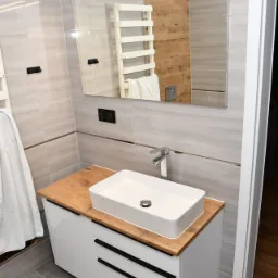 Nowoczesna łazienka z białą, prostokątną umywalką nablatową na szafce z drewnianym blatem i czarnymi uchwytami, szare płytki na ścianach i podłodze, lustro z oświetleniem LED, widoczny fragment...