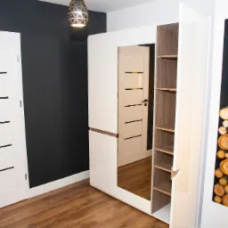 Biała szafa z lustrem i p&oacute;łkami, obok białe drzwi z czarnymi akcentami na tle grafitowej ściany, drewniana podłoga i dekoracyjne drewno ułożone w pionowej wnęce szafy.