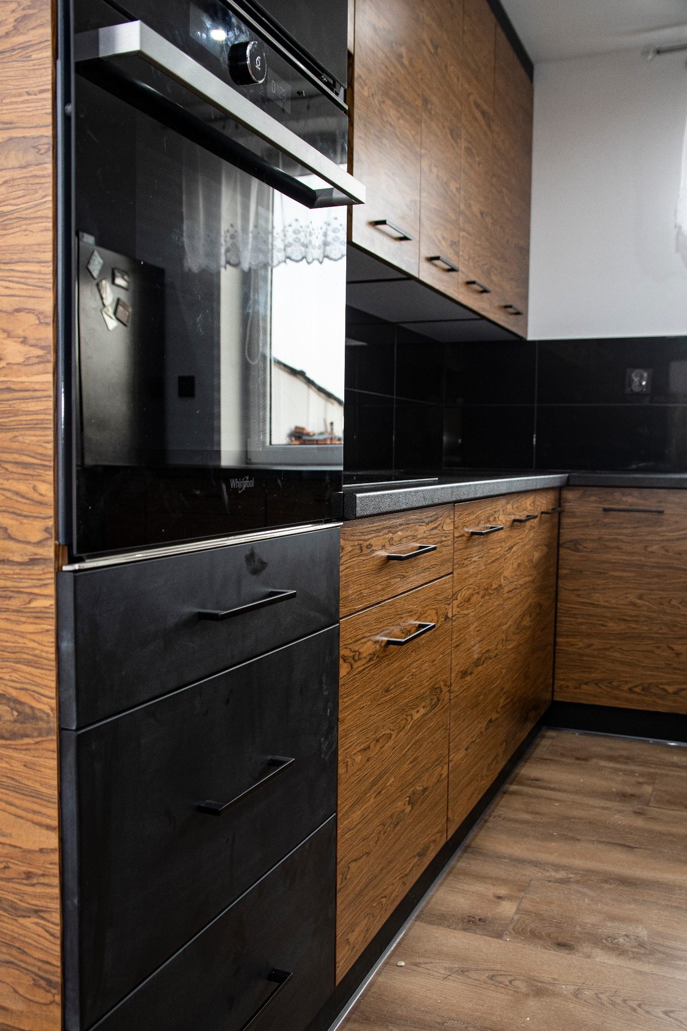 Nowoczesna kuchnia z fornirem w odcieniach brązu i czarnymi, matowymi frontami szuflad oraz piekarnikiem w zabudowie, czarny blat i płytki na ścianie, drewniana podłoga.