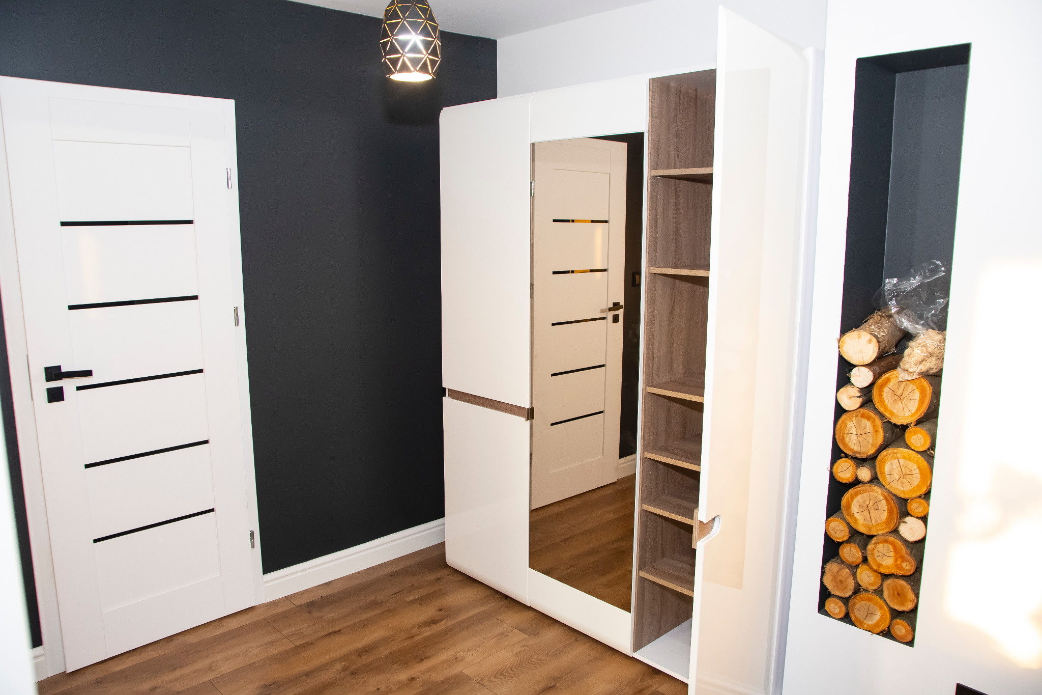 Biała szafa z lustrem i półkami, obok białe drzwi z czarnymi akcentami na tle grafitowej ściany, drewniana podłoga i dekoracyjne drewno ułożone w pionowej wnęce szafy.