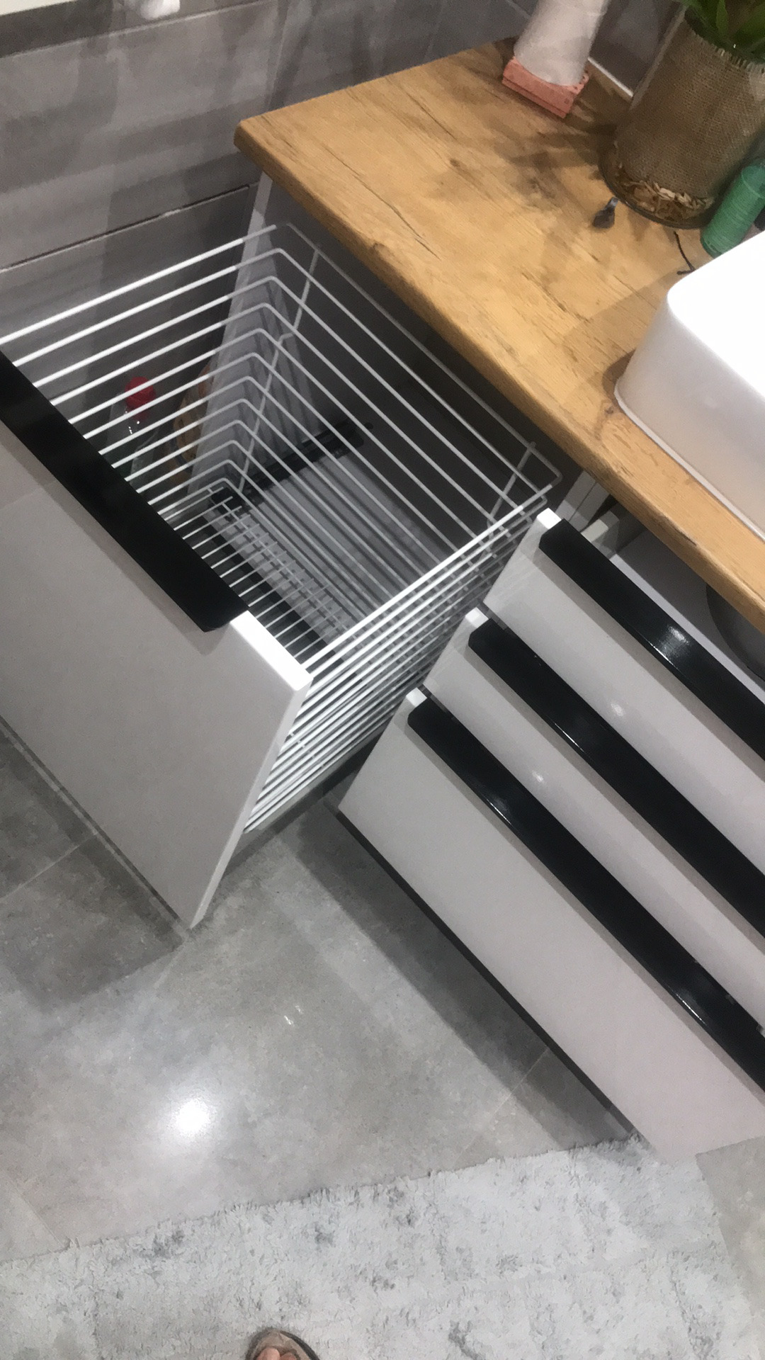 Wysuwana, druciana szuflada cargo w białej szafce łazienkowej z czarnymi akcentami, widok z góry, pod blatem z drewna.