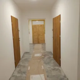 Korytarz w trakcie prac wykończeniowych: świeżo położona szara podłoga z płytek zabezpieczona kartonem, widoczne drzwi wejściowe po obu stronach i drewniane drzwi na końcu korytarza, ż&oacute;łta...