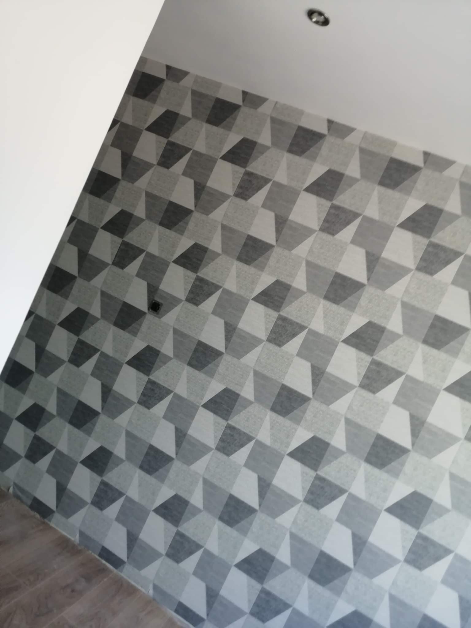 Szara ściana z geometrycznym wzorem 3D, widoczny fragment podłogi i sufit z zamontowanym oświetleniem punktowym.