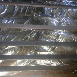 Izolacja poddasza folią aluminiową między drewnianymi belkami. Widoczne cegły w dolnej części kadru. Odbicie światła na folii tworzy unikalny wzór.