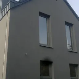 Elewacja budynku po tynkowaniu maszynowym: szary tynk, okna z szarymi ramami. Część budynku w trakcie prac wykończeniowych. Widok z dołu.