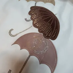 Biała tkanina z trzema naszytymi parasolkami w odcieniach beżu i brązu, każda ozdobiona błyszczącymi dżetami i drobnymi koralikami, tworzącymi wzory.