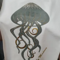 Biała koszulka z szarą aplikacją przedstawiającą meduzę ozdobioną złotymi i srebrnymi dżetami oraz złotym napisem 'by Madnes'.