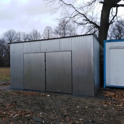 Korona - Metalowa szopa z dwuskrzydłowymi drzwiami przesuwnymi na terenie posesji w pochmurny dzień. Obok widoczny biały kontener z niebieskimi obramowaniami.