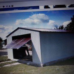 Korona - Metalowy garaż z otwartymi drzwiami w słoneczny dzień, widoczne trawnik i fragmenty nieba z chmurami.