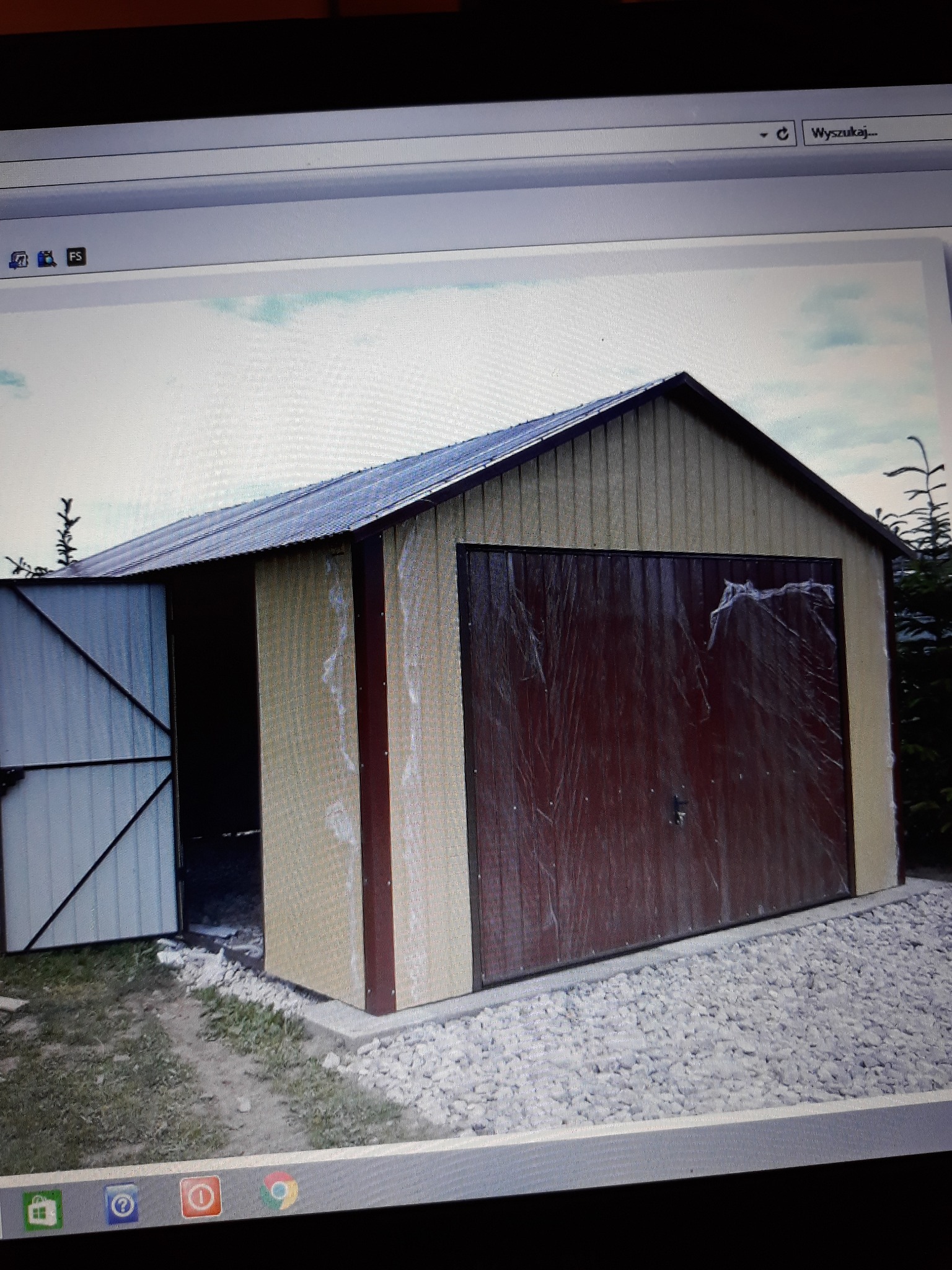 Nowy, blaszany garaż z uchylną bramą, częściowo jeszcze zabezpieczoną folią ochronną, położony na podłożu z białego grysu.