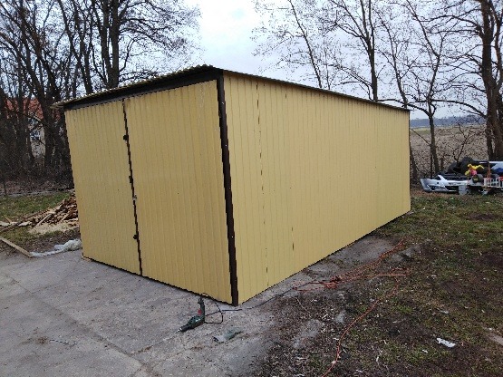 Żółty, blaszany garaż z dwuskrzydłowymi drzwiami na betonowym podłożu, z widocznymi narzędziami elektrycznymi i stertą drewna w tle, otoczony drzewami bez liści.