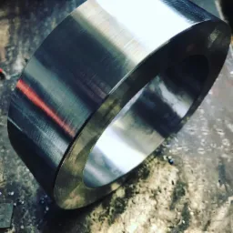 Metalowy pierścień po obr&oacute;bce tokarskiej, widoczne ślady obr&oacute;bki na powierzchni, leżący na zabrudzonym stole warsztatowym.