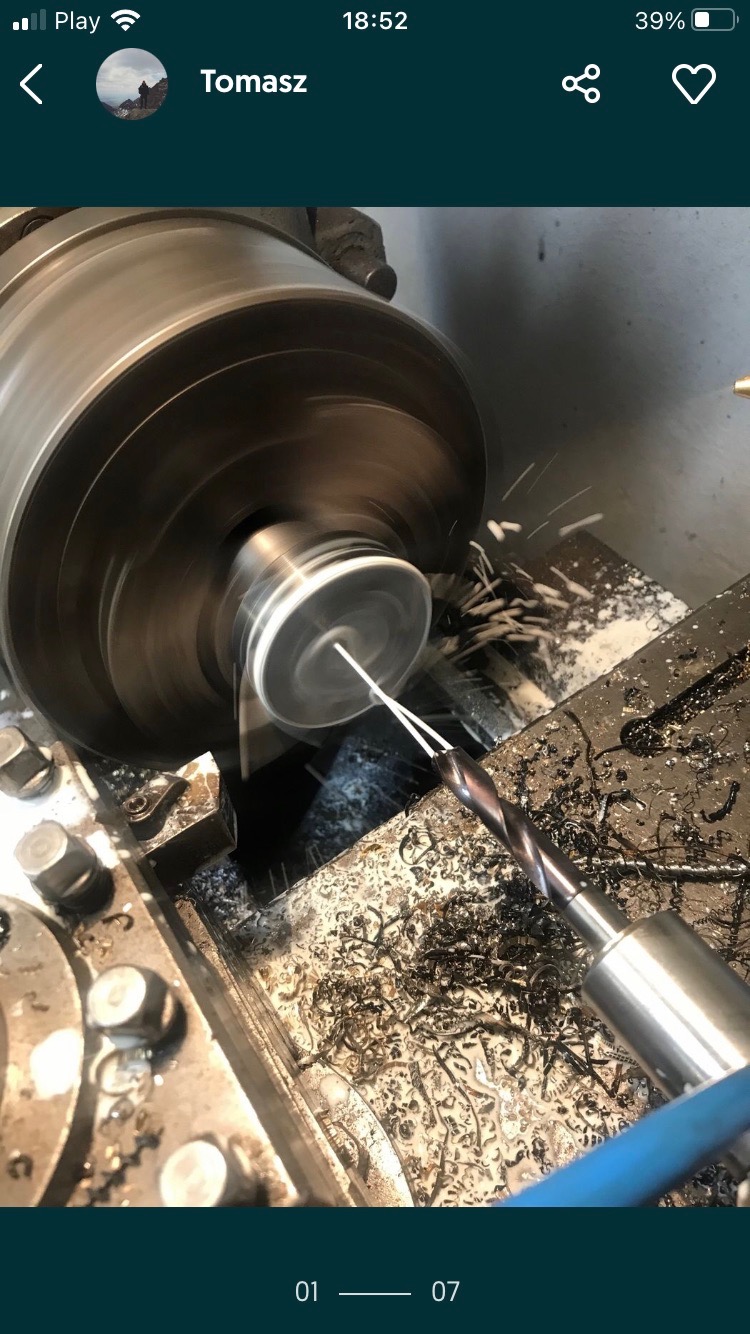 Metalowy element obrabiany w tokarce, wióry i chłodziwo w trakcie pracy, widoczny wiertło i uchwyt tokarski.