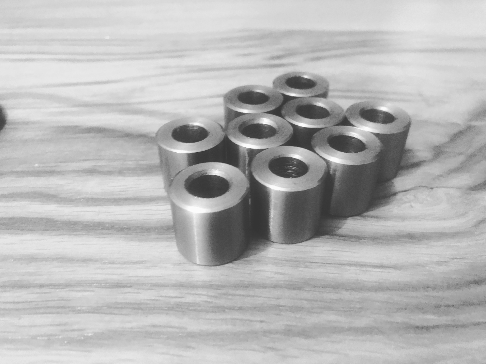Grupka ośmiu metalowych tulejek z gwintem wewnętrznym, ułożonych na drewnianej powierzchni w czarno-białej tonacji.