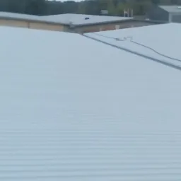 Widok z góry na nowo położone, jasnoszare, blaszane pokrycie dachowe z widocznymi liniami łączeń, na tle zabudowań i drzew w pochmurny dzień.