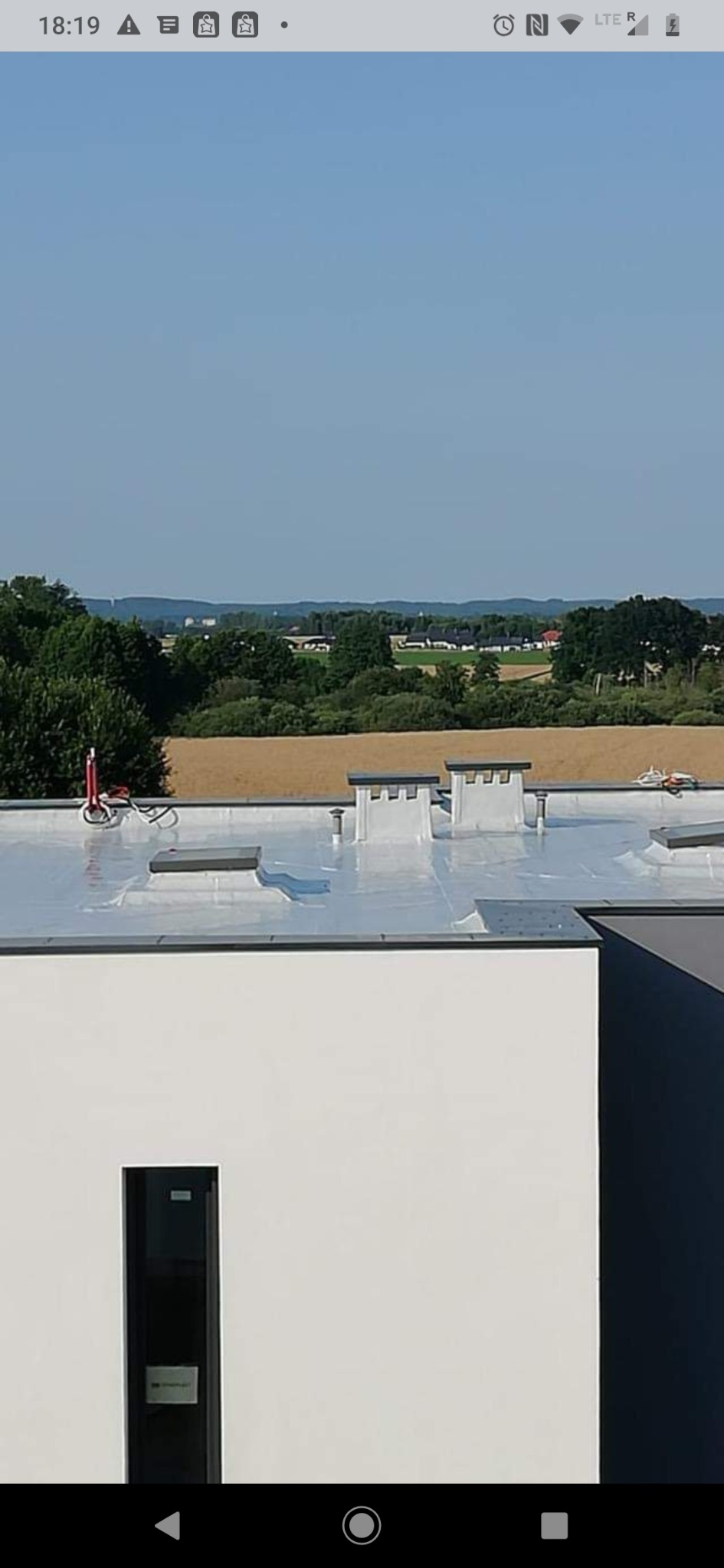 Widok z góry na płaski, srebrny dach z trzema kominami wentylacyjnymi i świetlikiem; w tle pola i lasy pod błękitnym niebem.